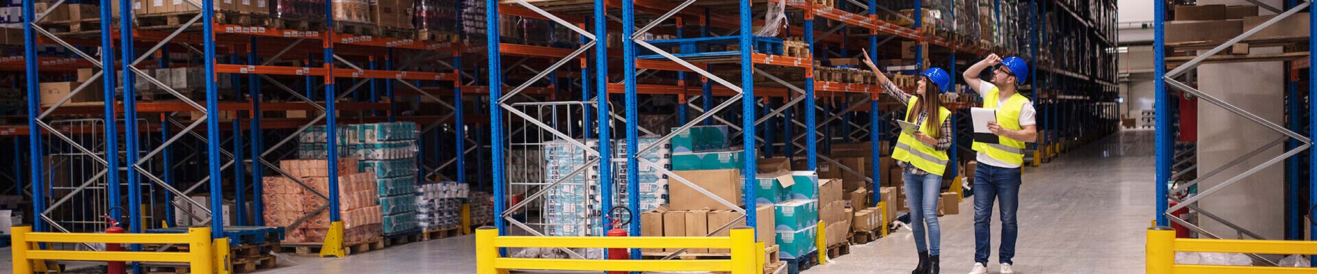 Two warehouse workers in blue helmets and yellow safety vests inspecting stocked shelves in a large industrial warehouse with roller conveyors in the foreground.
